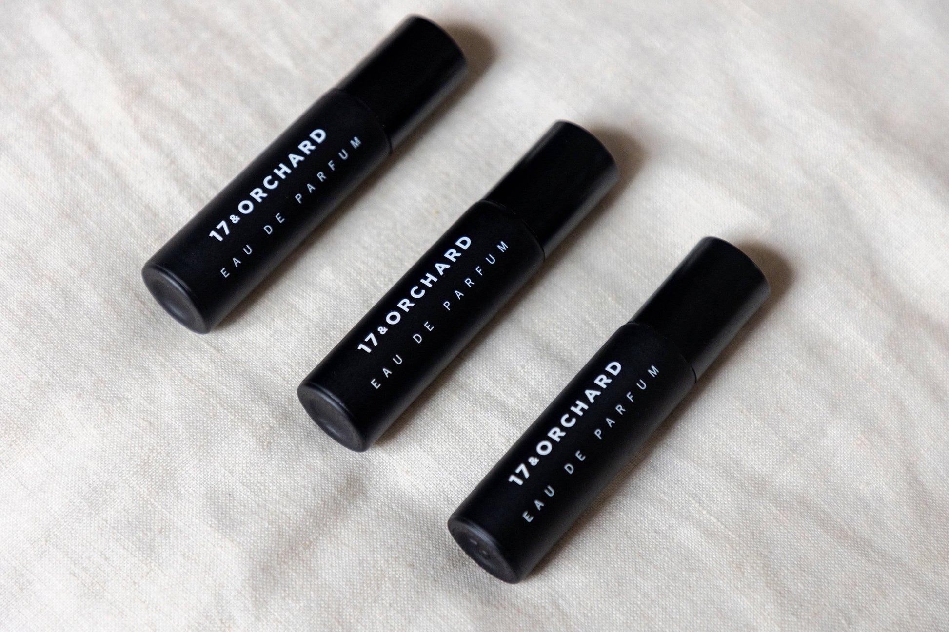 Three black perfume bottles labeled '17 Orchard Eau de Parfum' on a light fabric background.