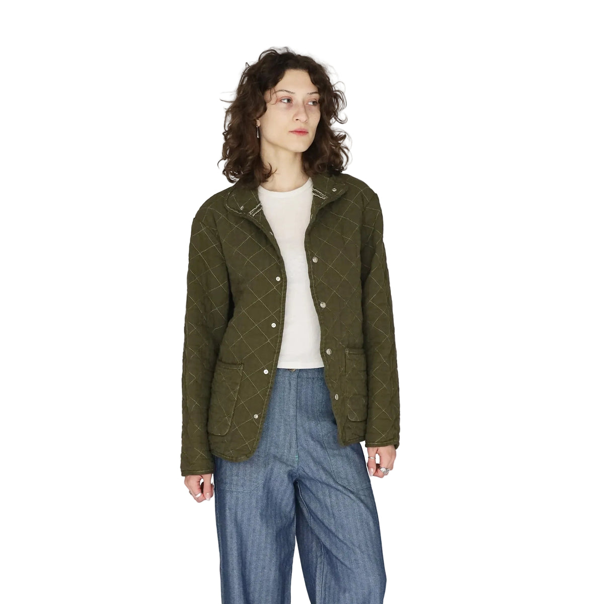A model wearing an olive green quilted jacket with a diamond pattern, styled over a white top and blue jeans, standing against a plain light gray background.