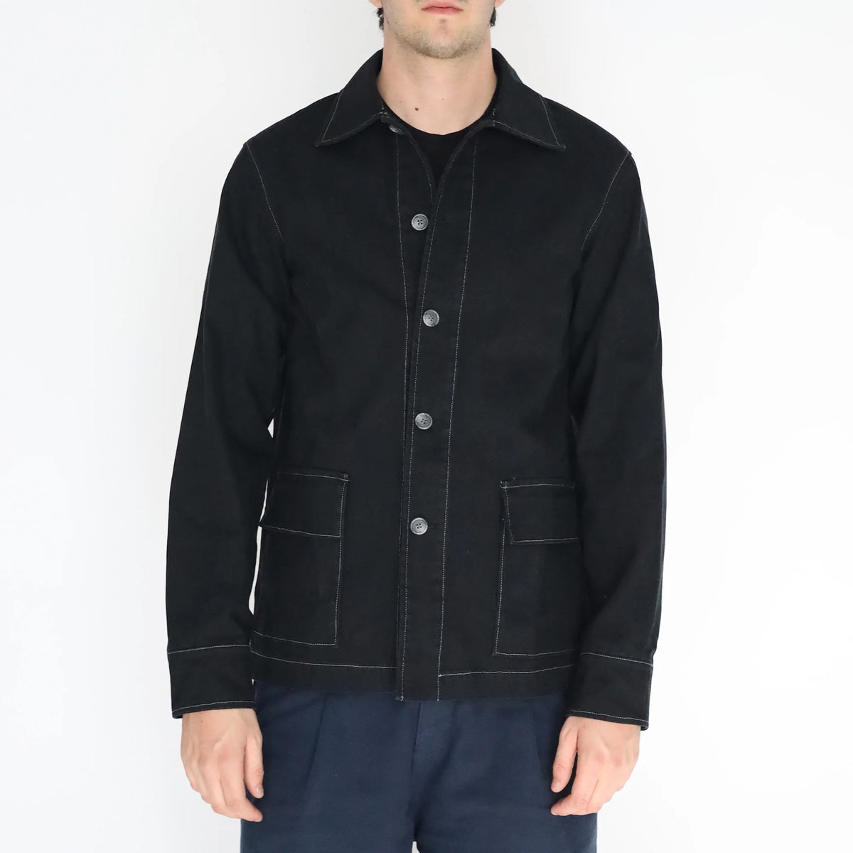A man wearing a black long-sleeve button-up jacket with white contrast stitching and silver buttons, paired with dark blue trousers, shot against a plain white background.