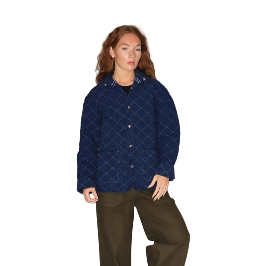 A model wearing a navy blue quilted jacket with a white grid pattern, paired with olive green pants, standing against a plain gray studio background.